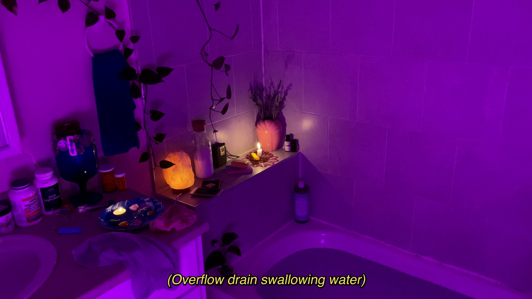 <span class='caption-artist'>Stefana Fratila</span>, <span class='caption-title'>Portrait of the Young (Crip) Artist at Bathtime</span>, <span class='caption-year'>2021</span>, <span class='caption-medium'>Video still</span>, <span class='caption-dimensions'>Image Description: In this film still, a purple light casts over the corner of a bathtub. To the left of the bathtub is a vanity. Various medication bottles line the back of the vanity to the right of the sink. On the corner of the vanity there is a blue plate with a lit tealight candle, a lighter, and towels. On the ledge behind the bathtub sits a glowing salt lamp, a lit candle, and other small treasures, as well as an illuminated iPhone. Above the ledge, two vines fall down against the tiled wall. The captions read, “Water pouring into bathtub.”</span>