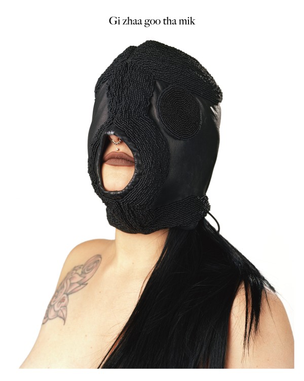 Dayna Danger, Gi zhaa goo tha mik, Full Moon Tibikigiizhiswabigoon Ikwe, 2017. Image Description: Photograph of a woman with long black straight hair draped over her left shoulder and a tattoo on her right shoulder. Her head and face are covered by a a black beaded BDSM mask. The mask has one opening revealing her lips, a nose piercing, and upper lip piercing.