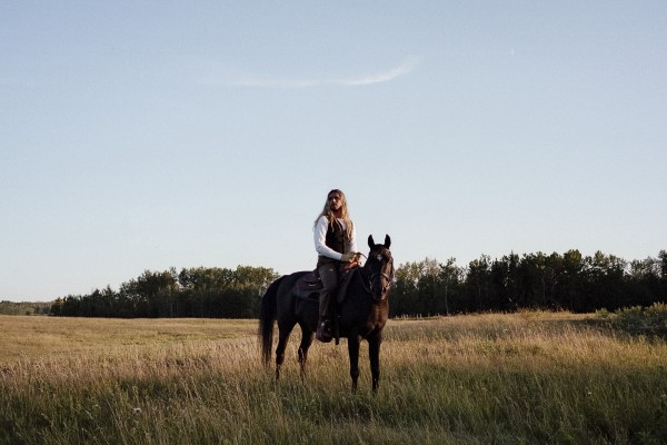 Vivek Shraya, Legends of the Trans, 2021. Image Description: Photograph of a figure on a horse in the middle of a grassy landscape with a forest and clear blue sky in the background. The figure has long blond hair, looks off into the distance, and is wearing a white dress shirt and a brown vest.