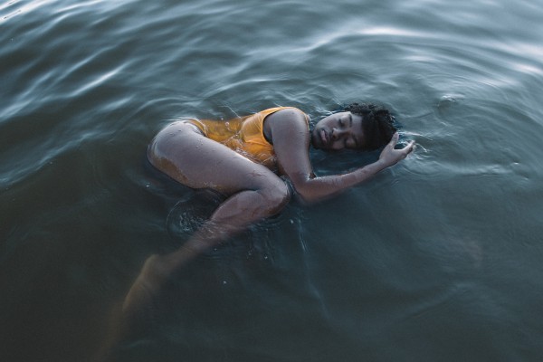 Gaëlle Elma, Harissa, 2018. Image Description: Photograph of a Black woman floating in a body of water. She is wearing a yellow bathing suit and is on her side with her knee and elbow bent. Her eyes are closed and she appears to be in a meditative state.