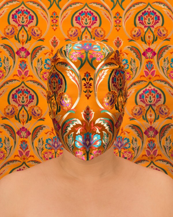 Chun Hua Catherine Dong, Skin Deep, 2019. Image Description: Photograph of a person directly facing the camera. The only part of their body that is revealed it their neck and shoulders. Their face is covered by a mask that matches the ornate pattern in the background of the photo. It is an orange Chinese silk design with pink, gold, yellow, and blue floral patterns.