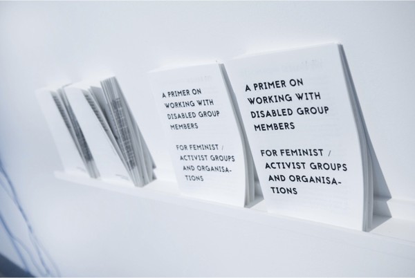 RA Walden, A Primer on Working With Disabled Group Members for Feminist / Activist Groups and Organisations, 2018 // Image description: A painted white shelf roughly 2 in/6 cm deep is mounted to a white wall. The photograph is taken at a slightly shifted perspective from the right side of the shelf, facing the left side. On the shelf are 4 stacks of printed pamphlets, leaning on the wall. The two left-hand side print-outs are facing backwards, and the two right-hand side printouts are facing forwards, with the title page showing. The title is printed in all upper-case letters and in black ink.
