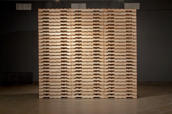Soheila Esfahani, My Place is Placeless, laser-etched wooden pallets, 10’ x 12’x 40” , 2013. Description: Three stacks of wooden pallets stand at around 12 feet tall. On the side face, the pallets are laser-engraved with a design that is Persianate in nature.